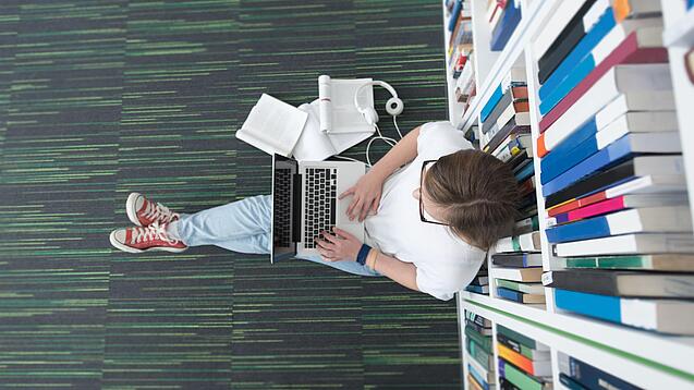 Junger Mensch sitzt mit einem Laptop auf dem Schoß vor einem Bücherregal auf dem Boden.