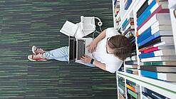 Junger Mensch sitzt mit einem Laptop auf dem Schoß vor einem Bücherregal auf dem Boden.