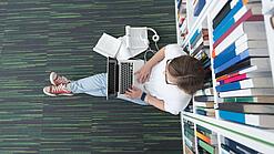 Junger Mensch sitzt mit einem Laptop auf dem Schoß vor einem Bücherregal auf dem Boden.