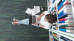 Junger Mensch sitzt mit einem Laptop auf dem Schoß vor einem Bücherregal auf dem Boden.