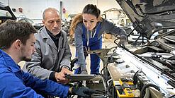 Zwei jüngere Menschen und ein älterer Mann sitzen in einer Autowerkstatt vor einer geöffneten Motorhaube.