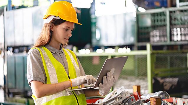 Junge Frau mit Schutzhelm, Warnweste und Arbeitshandschuhen an einem Laptop in einer Werkstatt