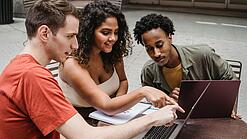 Drei junge Menschen sitzen um einen Laptop herum und schauen auf den Bildschirm.