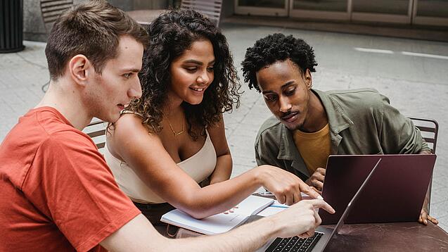 Drei junge Menschen sitzen um einen Laptop herum und schauen auf den Bildschirm.