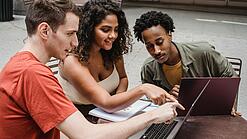 Drei junge Menschen sitzen um einen Laptop herum und schauen auf den Bildschirm.