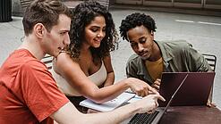 Drei junge Menschen sitzen um einen Laptop herum und schauen auf den Bildschirm.