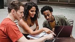Drei junge Menschen sitzen um einen Laptop herum und schauen auf den Bildschirm.
