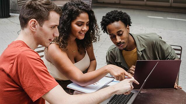 Drei junge Menschen sitzen um einen Laptop herum und schauen auf den Bildschirm.