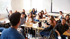 Eine Gruppe von Schülern sitzt in einem Klassenraum, der Lehrer steht vorne.