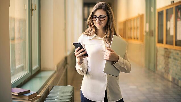 Eine Frau schaut auf ihr Handy, sie trägt einen Laptop unter dem Arm.