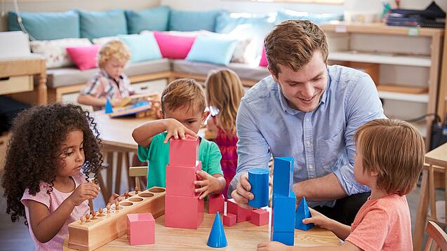 Ein Erzieher mit drei Kindern an einem Tisch, sie spielen mit Bauklötzen.