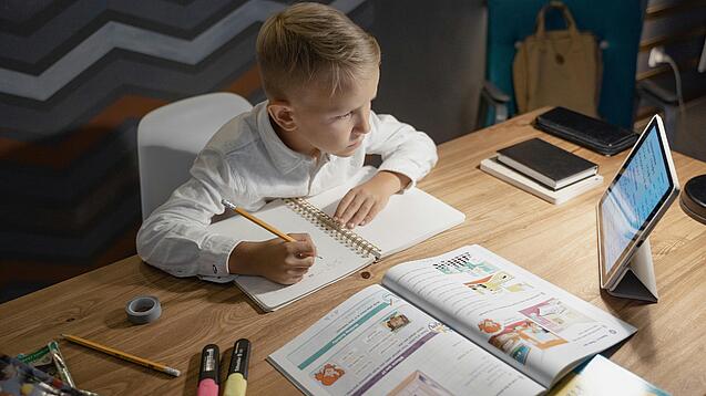 Junge sitzt an einem Schreibtisch und macht Schularbeiten mit einem Tablet.