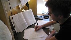Ein junger Mann sitzt an einem Schreibtisch und bearbeitet eine Aufgabe aus einem Schulbuch.