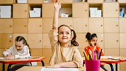 Ein junges Mädchen sitzt an einem Schulpult und meldet sich sehr motiviert, im Hintergrund sieht man weitere Schülerinnen an Tischen.