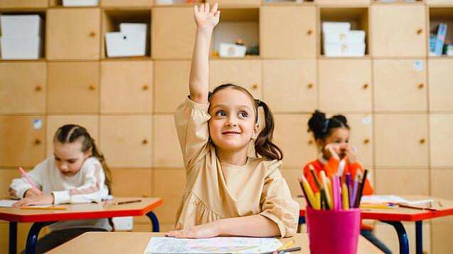 Ein junges Mädchen sitzt an einem Schulpult und meldet sich sehr motiviert, im Hintergrund sieht man weitere Schülerinnen an Tischen.