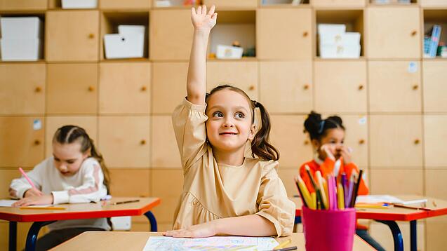 Ein junges Mädchen sitzt an einem Schulpult und meldet sich sehr motiviert, im Hintergrund sieht man weitere Schülerinnen an Tischen.