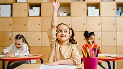 Ein junges Mädchen sitzt an einem Schulpult und meldet sich sehr motiviert, im Hintergrund sieht man weitere Schülerinnen an Tischen.