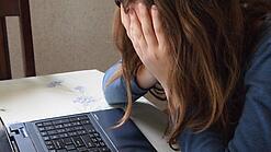 Eine junge Frau sitzt an einem Schreibtisch vor einem Laptop und hält sich die Hände vor das Gesicht.
