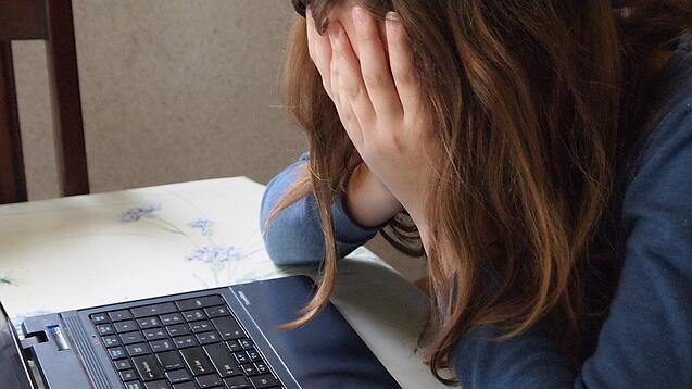 Eine junge Frau sitzt an einem Schreibtisch vor einem Laptop und hält sich die Hände vor das Gesicht.