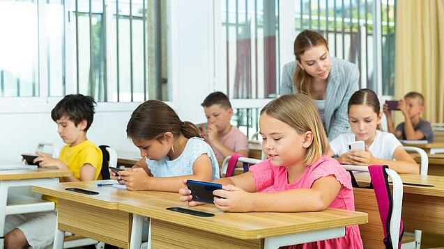 Mehrere Kinder sitzen an ihren Tischen im Klassenzimmer und schauen auf ihr jeweiliges Handy. Die Lehrerin schaut einer Schülerin über die Schulter.