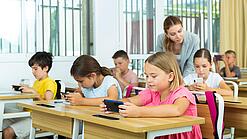 Mehrere Kinder sitzen an ihren Tischen im Klassenzimmer und schauen auf ihr jeweiliges Handy. Die Lehrerin schaut einer Schülerin über die Schulter.