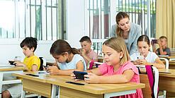 Mehrere Kinder sitzen an ihren Tischen im Klassenzimmer und schauen auf ihr jeweiliges Handy. Die Lehrerin schaut einer Schülerin über die Schulter.