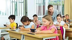 Mehrere Kinder sitzen an ihren Tischen im Klassenzimmer und schauen auf ihr jeweiliges Handy. Die Lehrerin schaut einer Schülerin über die Schulter.