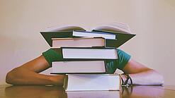Ein Mensch sitzt hinter einem Stapel Bücher und ist dadurch nicht weiter erkennbar.