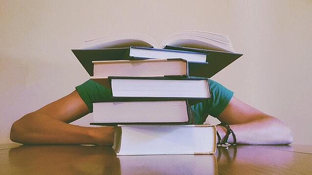 Ein Mensch sitzt hinter einem Stapel Bücher und ist dadurch nicht weiter erkennbar.
