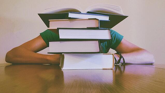 Ein Mensch sitzt hinter einem Stapel Bücher und ist dadurch nicht weiter erkennbar.