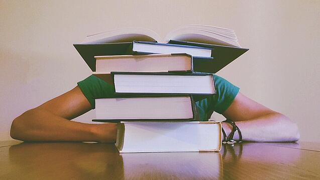 Ein Mensch sitzt hinter einem Stapel Bücher und ist dadurch nicht weiter erkennbar.