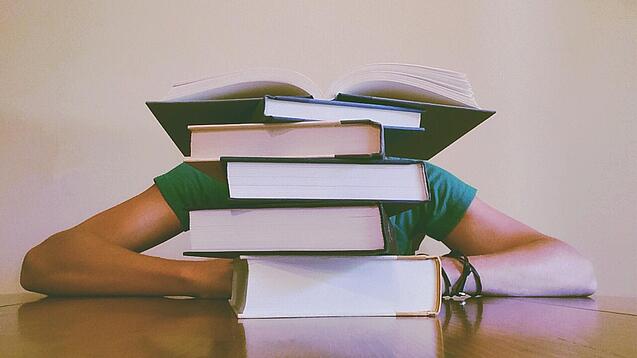 Ein Mensch sitzt hinter einem Stapel Bücher und ist dadurch nicht weiter erkennbar.