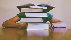 Ein Mensch sitzt hinter einem Stapel Bücher und ist dadurch nicht weiter erkennbar.