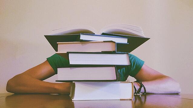 Ein Mensch sitzt hinter einem Stapel Bücher und ist dadurch nicht weiter erkennbar.
