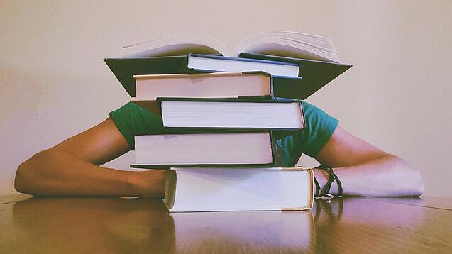 Ein Mensch sitzt hinter einem Stapel Bücher und ist dadurch nicht weiter erkennbar.