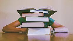 Ein Mensch sitzt hinter einem Stapel Bücher und ist dadurch nicht weiter erkennbar.