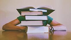 Ein Mensch sitzt hinter einem Stapel Bücher und ist dadurch nicht weiter erkennbar.