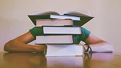 Ein Mensch sitzt hinter einem Stapel Bücher und ist dadurch nicht weiter erkennbar.