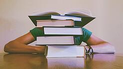 Ein Mensch sitzt hinter einem Stapel Bücher und ist dadurch nicht weiter erkennbar.