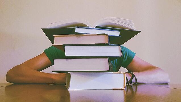Ein Mensch sitzt hinter einem Stapel Bücher und ist dadurch nicht weiter erkennbar.
