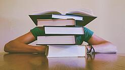 Ein Mensch sitzt hinter einem Stapel Bücher und ist dadurch nicht weiter erkennbar.