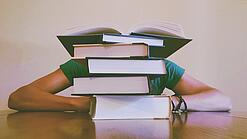 Ein Mensch sitzt hinter einem Stapel Bücher und ist dadurch nicht weiter erkennbar.