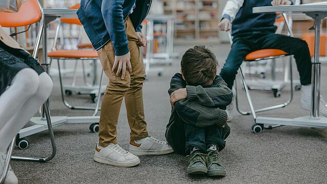 Kleiner Junge sitzt am Boden und verbirgt Gesicht während andere Kinder lachend auf ihn zeigen.