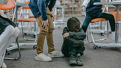 Kleiner Junge sitzt am Boden und verbirgt Gesicht während andere Kinder lachend auf ihn zeigen.