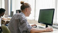 Ein junger Mann sitzt an einem Schreibtisch in einem Klassenzimmer und arbeitet an einem Computer.