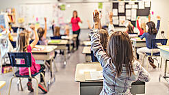 Grundschulkinder sitzen an ihren Tischen und schauen auf eine Lehrerin.