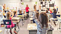 Grundschulkinder sitzen an ihren Tischen und schauen auf eine Lehrerin.