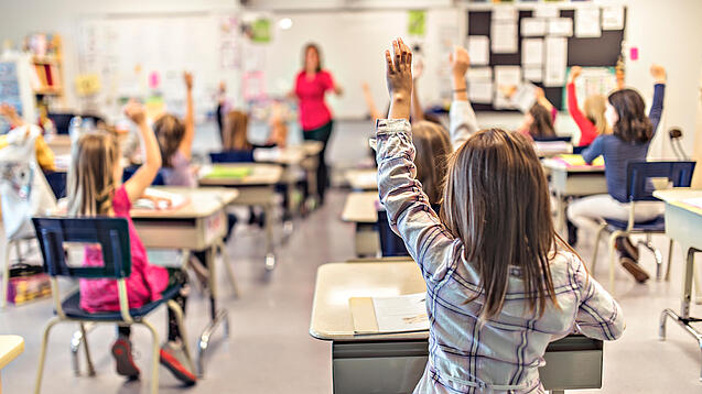 Grundschulkinder sitzen an ihren Tischen und schauen auf eine Lehrerin.