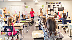 Grundschulkinder sitzen an ihren Tischen und schauen auf eine Lehrerin.