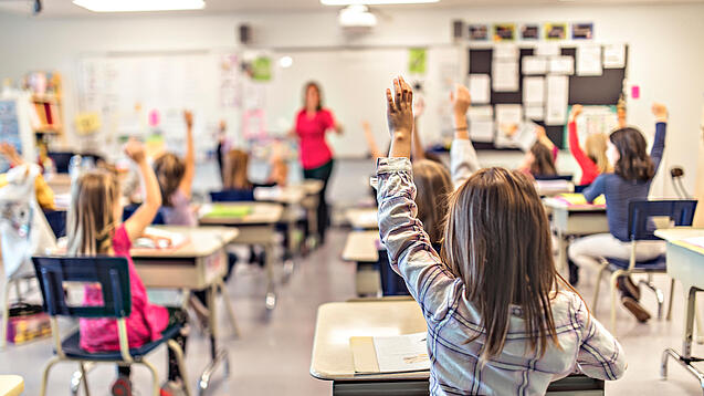Grundschulkinder sitzen an ihren Tischen und schauen auf eine Lehrerin.